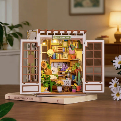 Wooden model of a store with various items on a wooden base, surrounded by plants and decor.