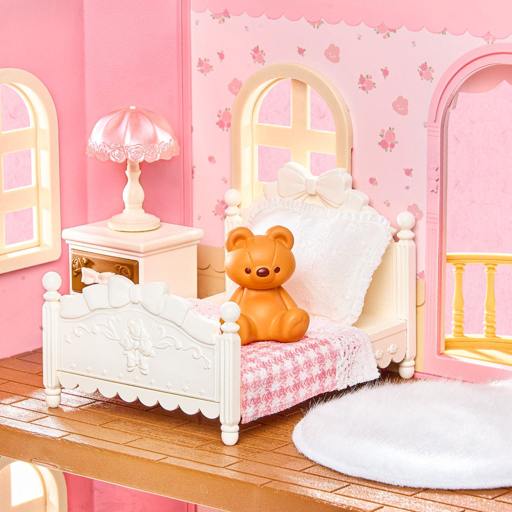 A dollhouse-style bedroom scene with a white bed, pink patterned bedding, a small orange teddy bear sitting on the bed, a pink nightstand with a lamp, and pink walls with floral wallpaper.