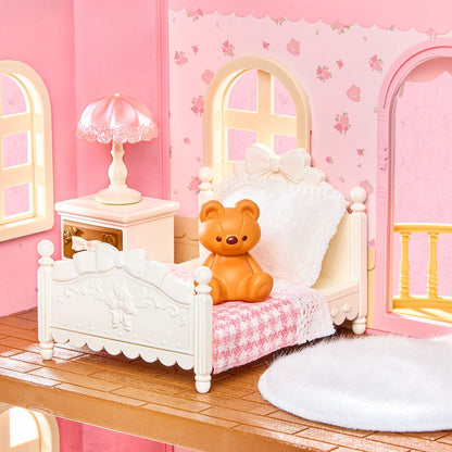 A dollhouse-style bedroom scene with a white bed, pink patterned bedding, a small orange teddy bear sitting on the bed, a pink nightstand with a lamp, and pink walls with floral wallpaper.
