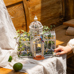A miniature white metal greenhouse model with a domed roof and floral details, displayed on a draped white cloth on a wooden table. A hand is reaching toward the small object, with limes and rustic wood in the background.