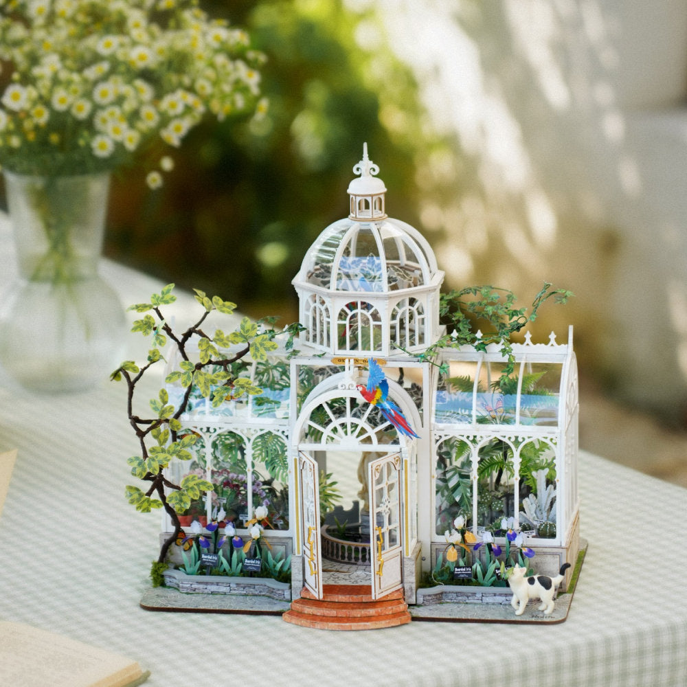 A miniature white greenhouse model with glass panels, small plants, flowers, a parrot figurine, and tiny animals, displayed on a table in a softly blurred outdoor setting.