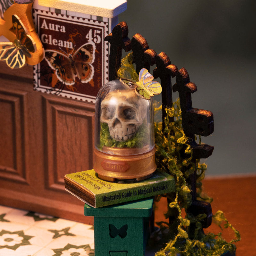 A miniature decorative display featuring a glass dome with a small skull and butterfly, placed on a green pedestal next to a dark wooden arch with yellow-green moss, in a styled tabletop scene.