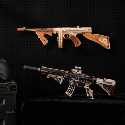 Two miniature toy or model rifles displayed against a black pegboard background, one mounted on the wall and one resting on a black surface below. The top rifle is tan and brown with a red tip, and the bottom rifle is black, tan, and white with a red tip.