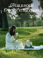 A woman in a white dress sits on a blanket in a grassy park, arranging small glass terrariums. Large white text at the top reads 'Build your own little oasis'.