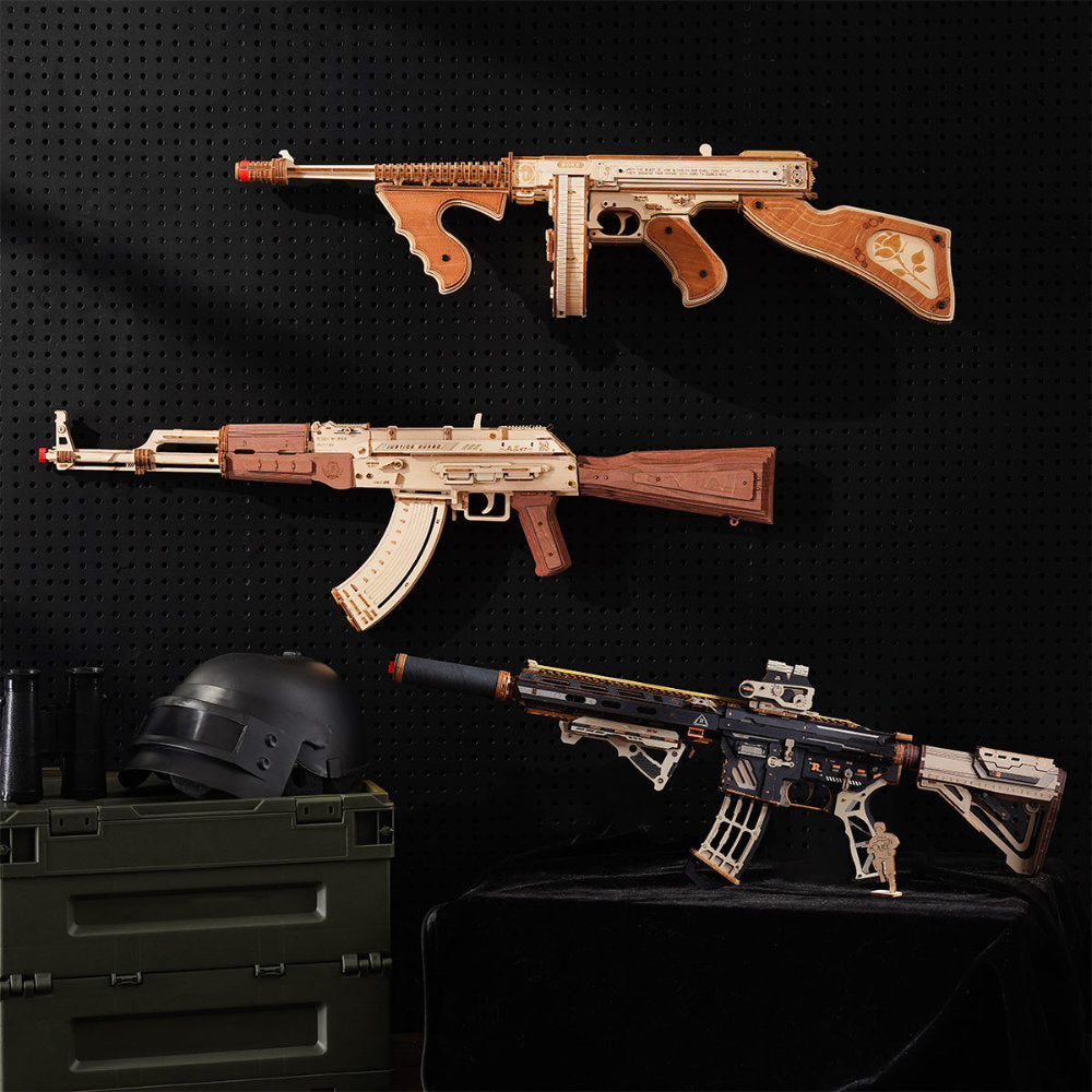Three decorative replica rifle wall pieces and a black tactical-style rifle displayed against a black pegboard wall, with a dark helmet and green storage crate as props.