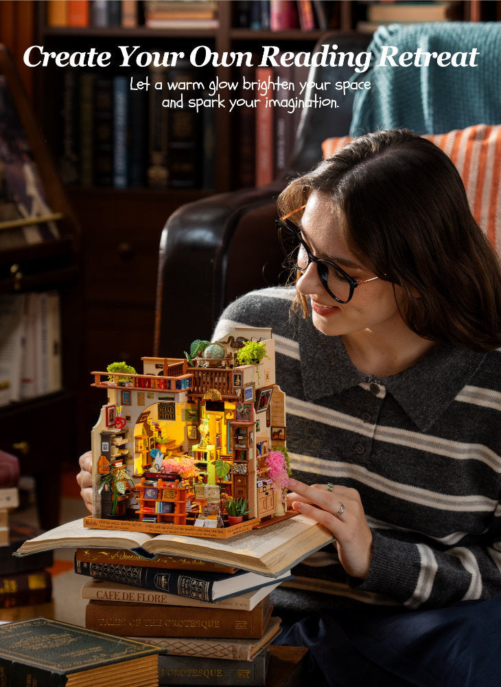 A woman in a striped sweater and glasses holds a miniature reading nook model built on top of open books in a cozy library setting. The scene includes text at the top reading 'Create Your Own Reading Retreat'.