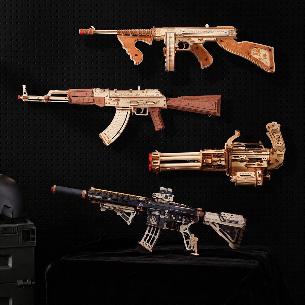 Four miniature toy or model rifles displayed against a black pegboard background, including a long rifle, an AK-style rifle, a minigun, and a black-and-silver assault rifle, with a helmet partially visible at lower left.