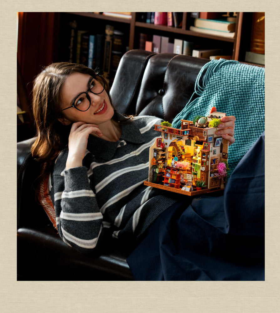 A woman wearing glasses and a striped sweater relaxes in a dark leather armchair, holding a small illuminated wooden house model with tiny colorful details. A teal knit blanket is draped over the chair, and bookshelves are visible in the background.