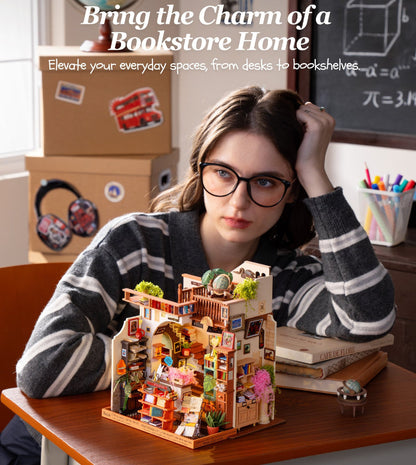 A woman with glasses sits at a wooden desk in a classroom-like setting, looking thoughtfully at a small, highly detailed miniature bookshelf diorama. The diorama features a cozy bookshop interior with miniature shelves, books, plants, and characters.