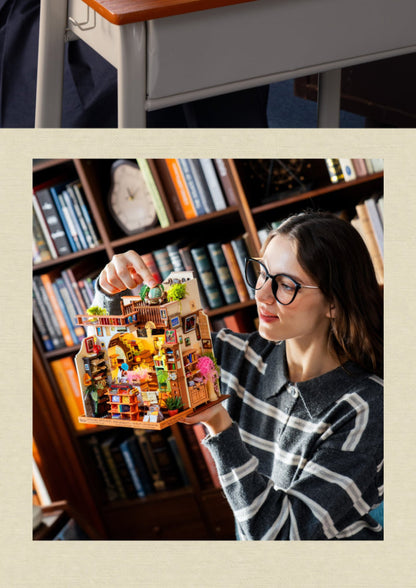 A woman wearing glasses holds a detailed miniature house model in front of a bookshelf. The top strip shows a close-up of a wooden desk surface.