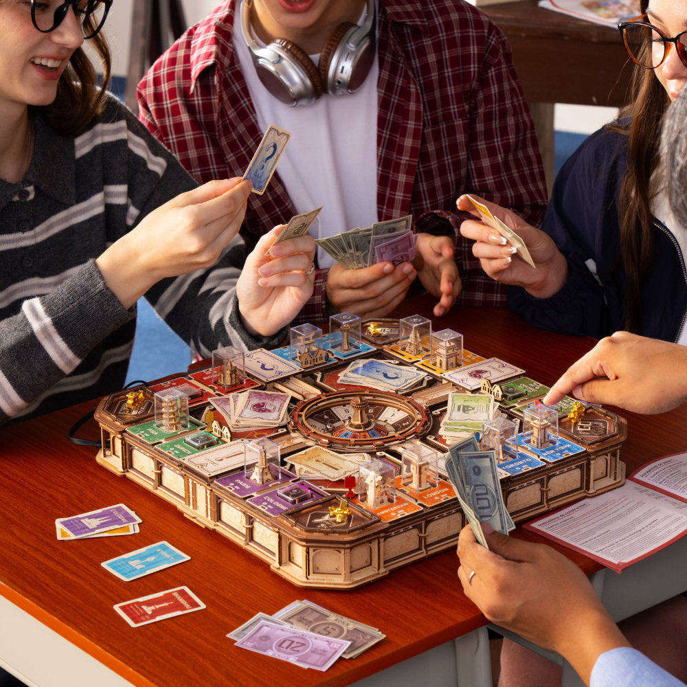 A group of people playing a detailed board game with money, cards, and miniature vehicles around a wooden table.