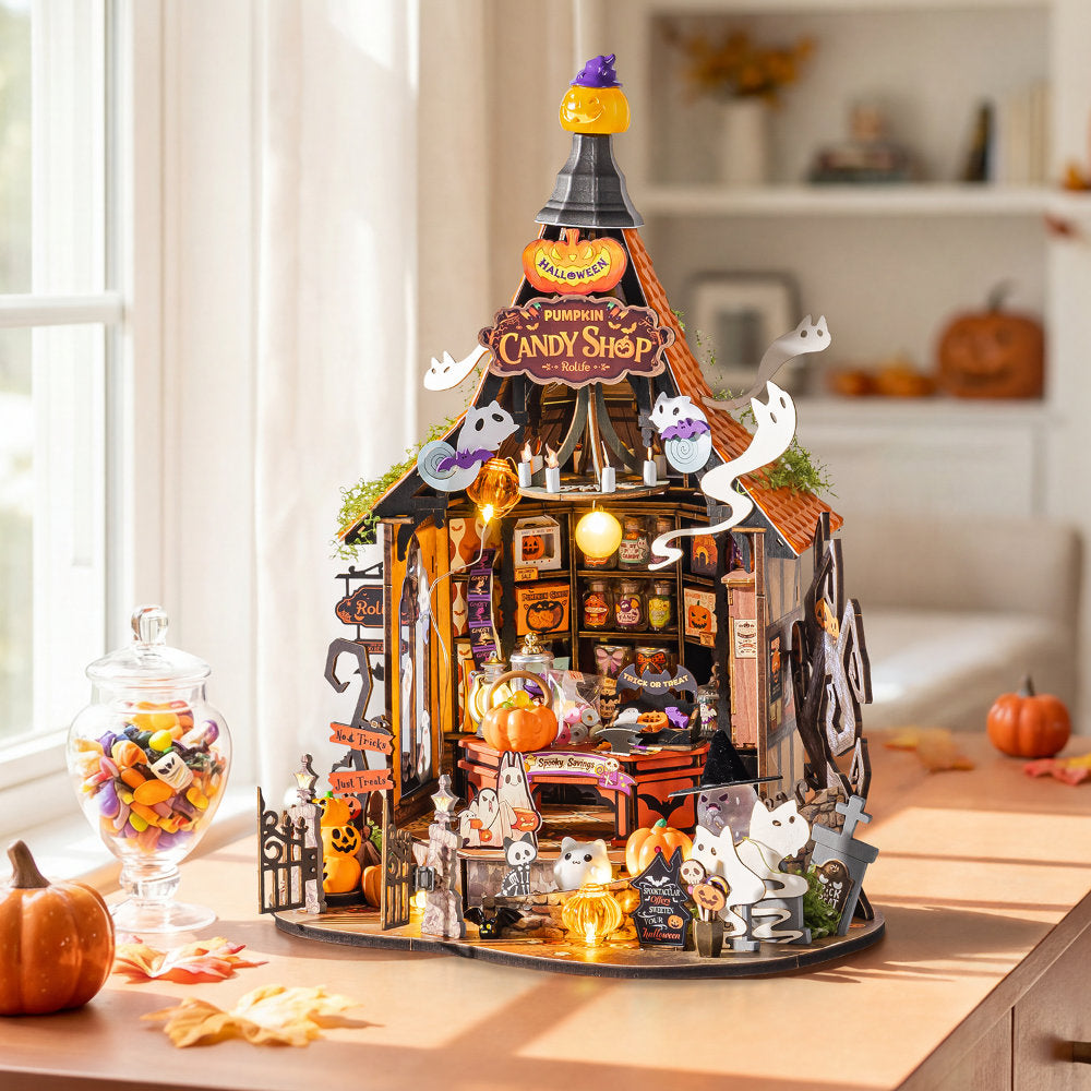 A detailed miniature Halloween diorama set up on a wooden table in a cozy, softly lit room. The set features a small 'Pumpkin Candy Shop' building with glowing orange windows, hanging jack-o'-lanterns, ghost decorations, miniature candy jars, and tiny figurines.