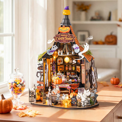 A detailed miniature Halloween diorama set up on a wooden table in a cozy, softly lit room. The set features a small 'Pumpkin Candy Shop' building with glowing orange windows, hanging jack-o'-lanterns, ghost decorations, miniature candy jars, and tiny figurines.