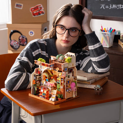 A young woman with glasses and a striped sweater sits at a desk, looking thoughtfully at a detailed miniature house model. The room has books, a chalkboard, and cardboard boxes in the background.