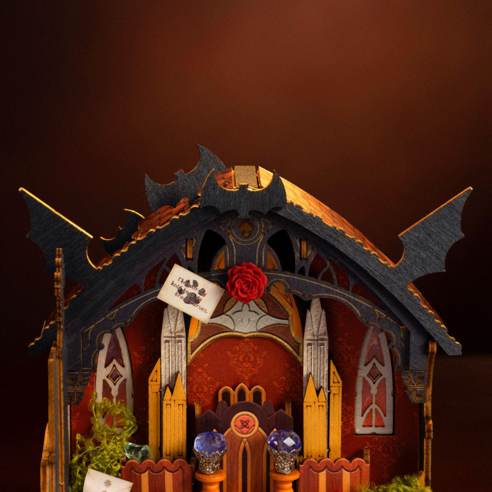 A miniature gothic cathedral or cathedral-like diorama model with bat-wing shaped roof spires, red and gold interior detailing, small windows, a red rose accent, and tiny decorative elements, shown against a dark brown gradient background.