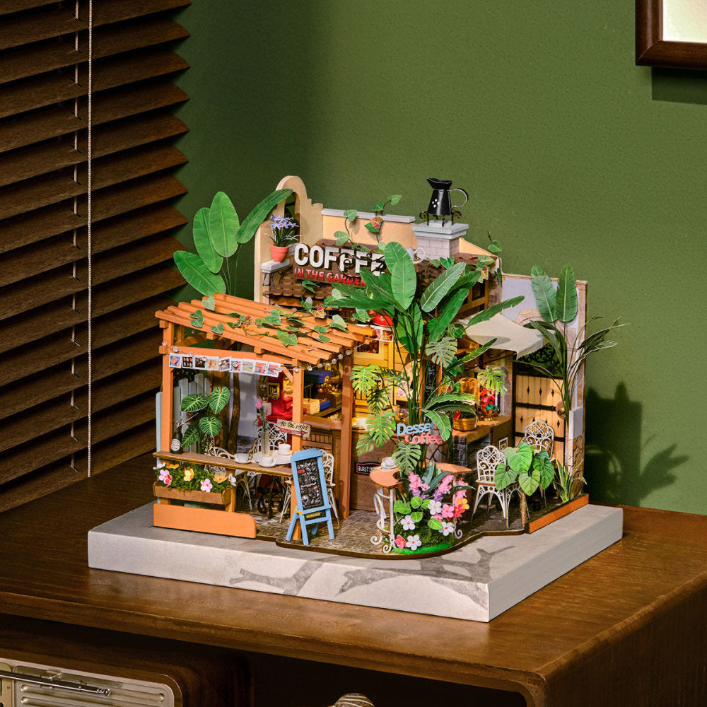 A miniature coffee shop dollhouse model displayed on a marble-look base, set on a wooden table against a green wall with window blinds in the background.
