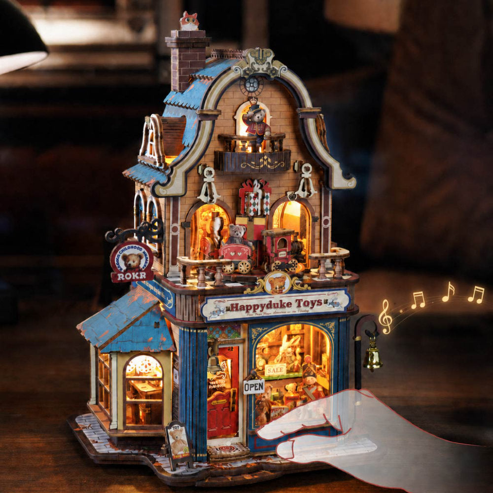 A miniature two-story toy shop figurine with warm LED lighting, detailed architectural features, and a hand interacting with it on a wooden surface.