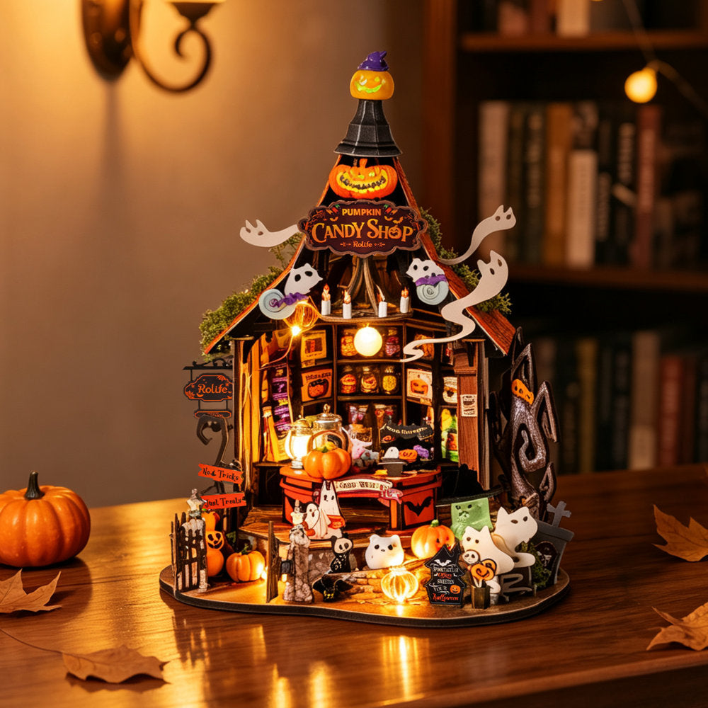 A detailed miniature Halloween diorama set up on a wooden table, featuring a lit 'Candy Shop' structure with ghosts, pumpkins, a witch, and small figures, photographed in a cozy indoor setting with warm ambient lighting.