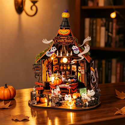 A detailed miniature Halloween diorama set up on a wooden table, featuring a lit 'Candy Shop' structure with ghosts, pumpkins, a witch, and small figures, photographed in a cozy indoor setting with warm ambient lighting.
