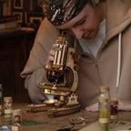 A man wearing a beige hoodie and a patterned headband peers closely through a wooden and brass compound microscope on a table, with small specimen jars in the blurred background.