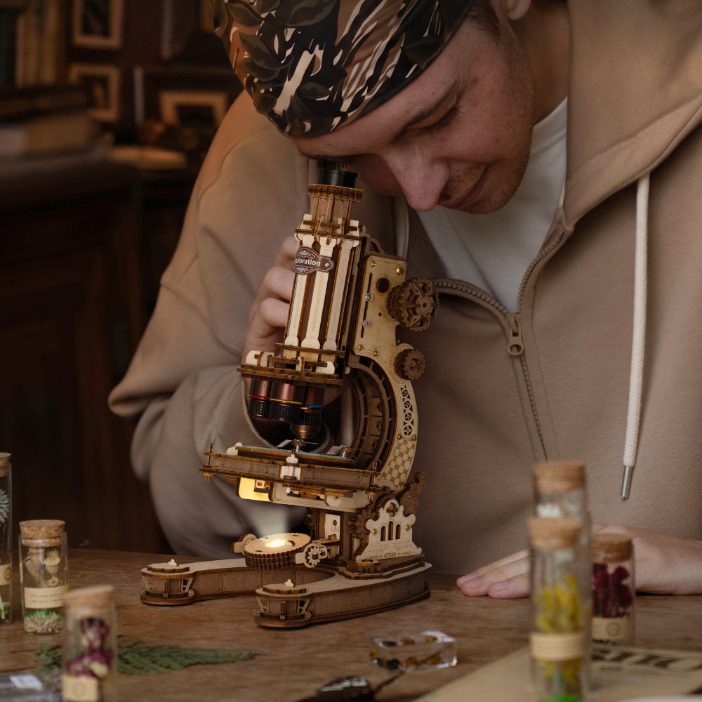 A man wearing a beige hoodie and a patterned headband peers closely through a wooden and brass compound microscope on a table, with small specimen jars in the blurred background.