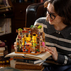 A woman wearing glasses and a gray striped sweater holds a miniature wooden cafe house model built on top of an open book. The detailed dollhouse-style structure has warm yellow interior lights, small green plants, and colorful decor, with books stacked underneath in a cozy indoor setting.