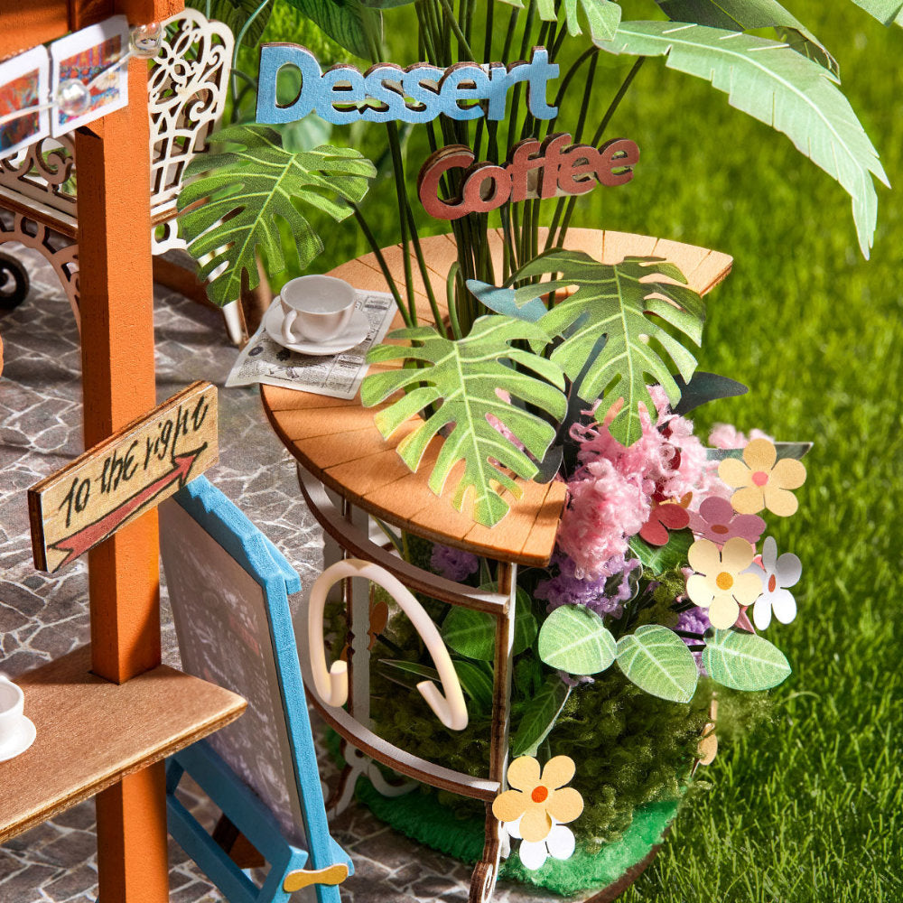 A miniature dollhouse-scale cafe scene with a small wooden table decorated with artificial monstera leaves, flowers, and a cup, alongside a sign reading 'Dessert Coffee' and a miniature chair.