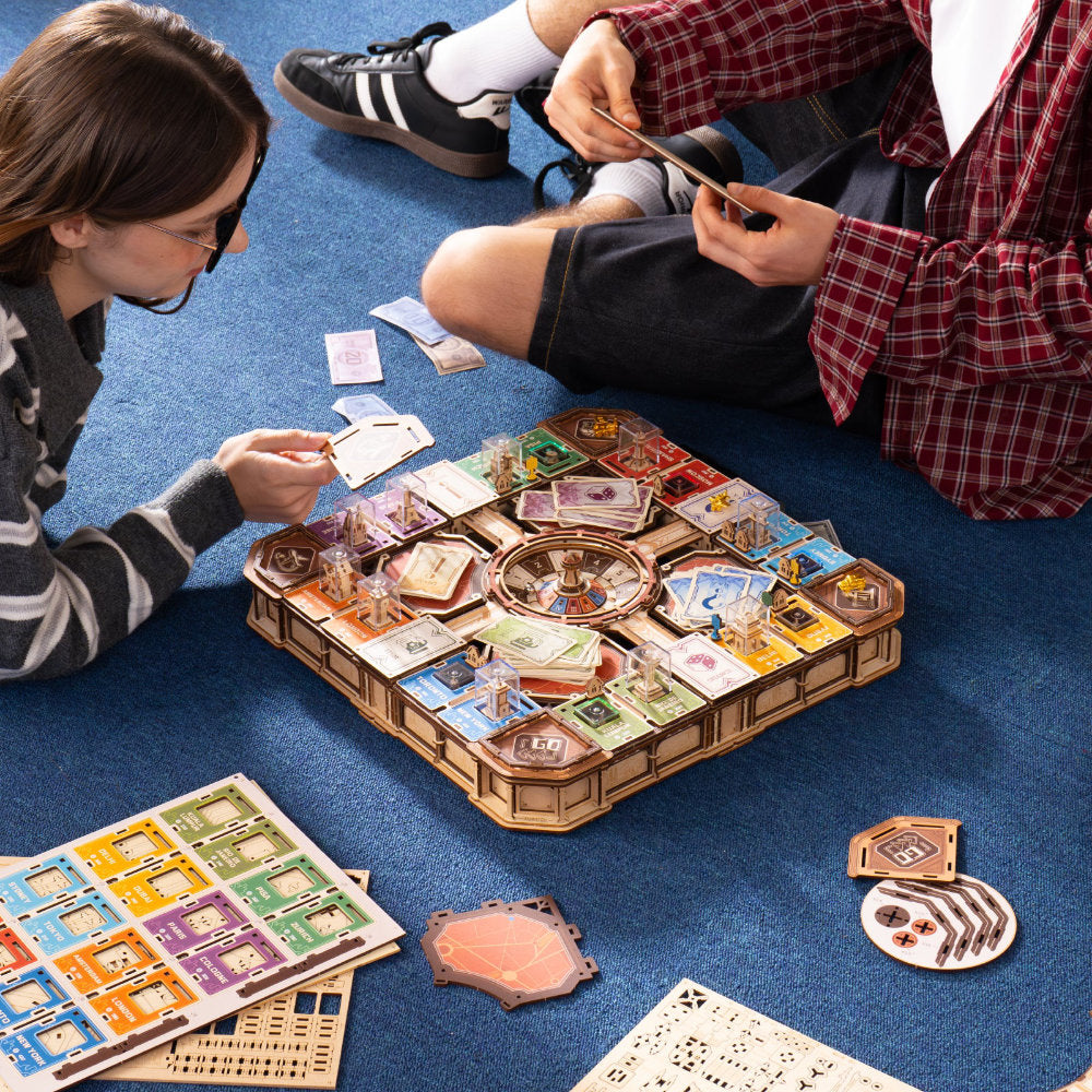 A group of people sitting on a blue carpet playing a board game with cards and game pieces spread out on the floor.