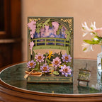 Decorative card with a pond scene featuring flowers and a bridge on a wooden table.