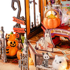A styled Halloween-themed miniature display with orange jack-o'-lantern decorations, a small pumpkin basket, ghost cutouts, a faux gate, and a lit white cat figurine on a tabletop.