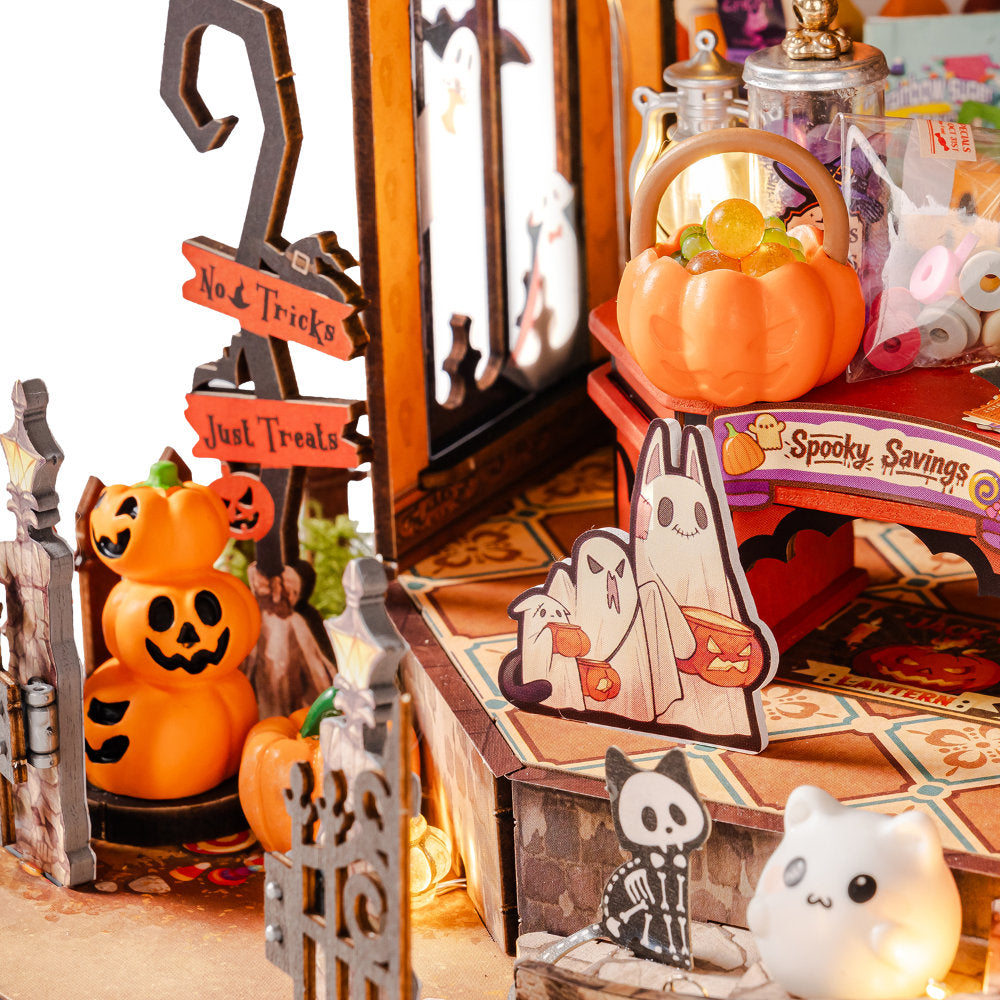 A styled Halloween-themed miniature display with orange jack-o'-lantern decorations, a small pumpkin basket, ghost cutouts, a faux gate, and a lit white cat figurine on a tabletop.