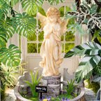 A cream-colored 'Water Lily' angel garden statue standing on a faux-stone base inside a circular planter with water lilies, surrounded by green tropical leaves in front of a white arched door.