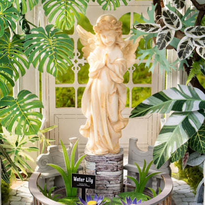 A cream-colored 'Water Lily' angel garden statue standing on a faux-stone base inside a circular planter with water lilies, surrounded by green tropical leaves in front of a white arched door.