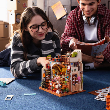 Two young people assembling a detailed miniature dollhouse in a room with cardboard boxes in the background. The dollhouse features a warm, cozy interior with tiny furniture pieces, plants, and shelves.