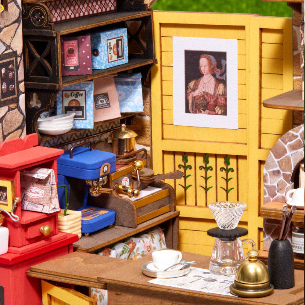 A miniature coffee shop scene with a yellow wooden counter, coffee maker, cups, shelves, and a framed portrait on a tiny tabletop.