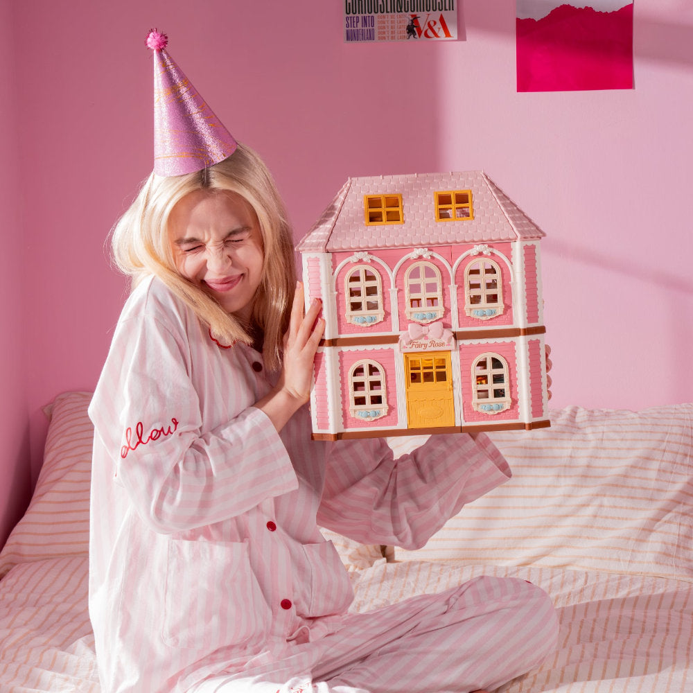 A woman in pink pajamas and a glittery party hat sits on a bed holding a pink dollhouse. The room has pink walls and striped bedding.