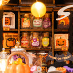 A Halloween-themed candy display with glass jars of assorted gummies and sweets on a wooden shelf, alongside pumpkin decorations, a hanging yellow balloon, and sale signs.