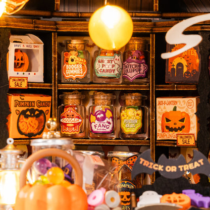 A Halloween-themed candy display with glass jars of assorted gummies and sweets on a wooden shelf, alongside pumpkin decorations, a hanging yellow balloon, and sale signs.