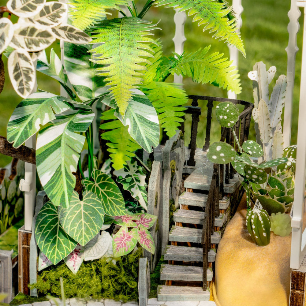 A miniature dollhouse scene featuring a small wooden staircase leading up a ramp, surrounded by detailed artificial plants, cacti, and a white picket fence.