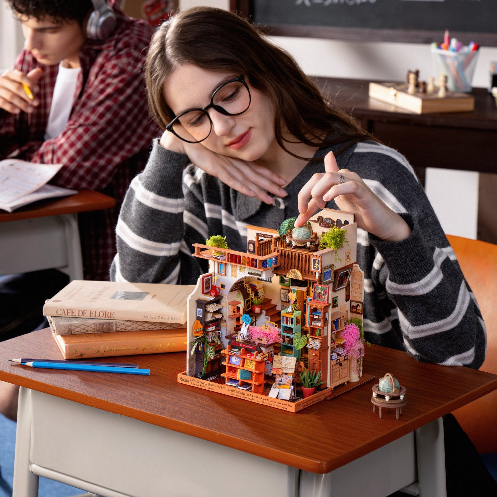 A woman in a classroom setting is building a detailed miniature dollhouse with tiny furniture and characters. The dollhouse features a warm orange-brown frame with colorful interior details, shelves, plants, and small figurines.