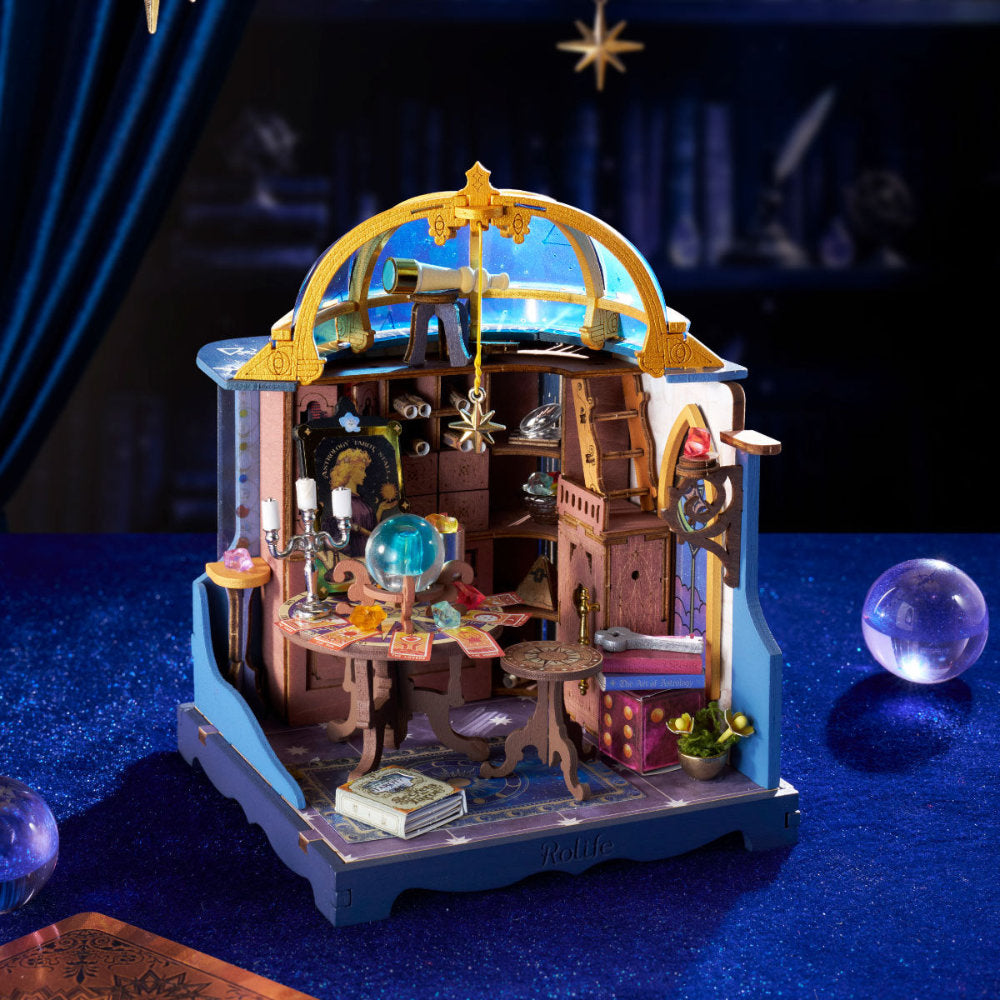 A miniature fantasy observatory and library dollhouse set on a dark blue textured surface, featuring a blue and gold dome, wooden interior shelves, a telescope, books, and small decorative props.