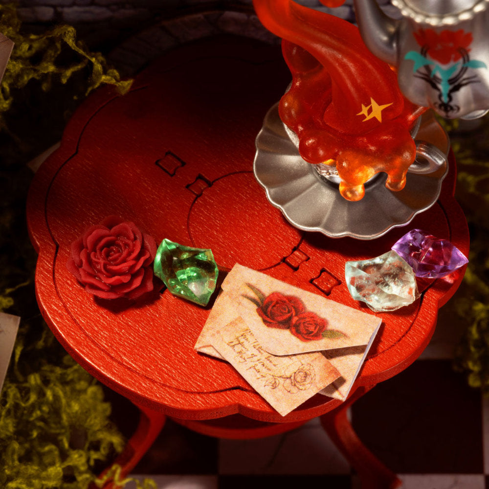 A styled tabletop scene with a red scalloped table holding a silver teapot with orange-red wax dripping over it, a red rose, green and purple faceted gemstones, and a vintage-style letter with a rose illustration.