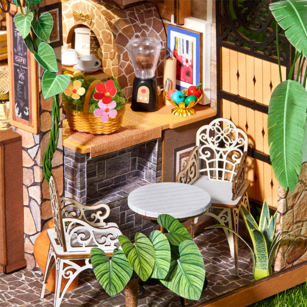 A miniature dollhouse-style cafe interior with white metal chairs, a small round table, a blender, flower baskets, stone walls, and tropical green plants.