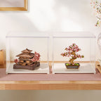 Two clear acrylic display boxes on a white surface, one containing a miniature traditional Japanese pagoda model with pink cherry blossoms, and the other containing a bonsai-style flowering tree in a rectangular pot.