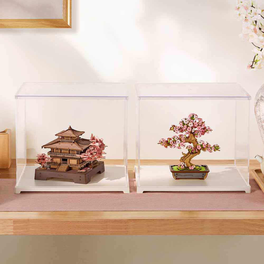 Two clear acrylic display boxes on a white surface, one containing a miniature traditional Japanese pagoda model with pink cherry blossoms, and the other containing a bonsai-style flowering tree in a rectangular pot.