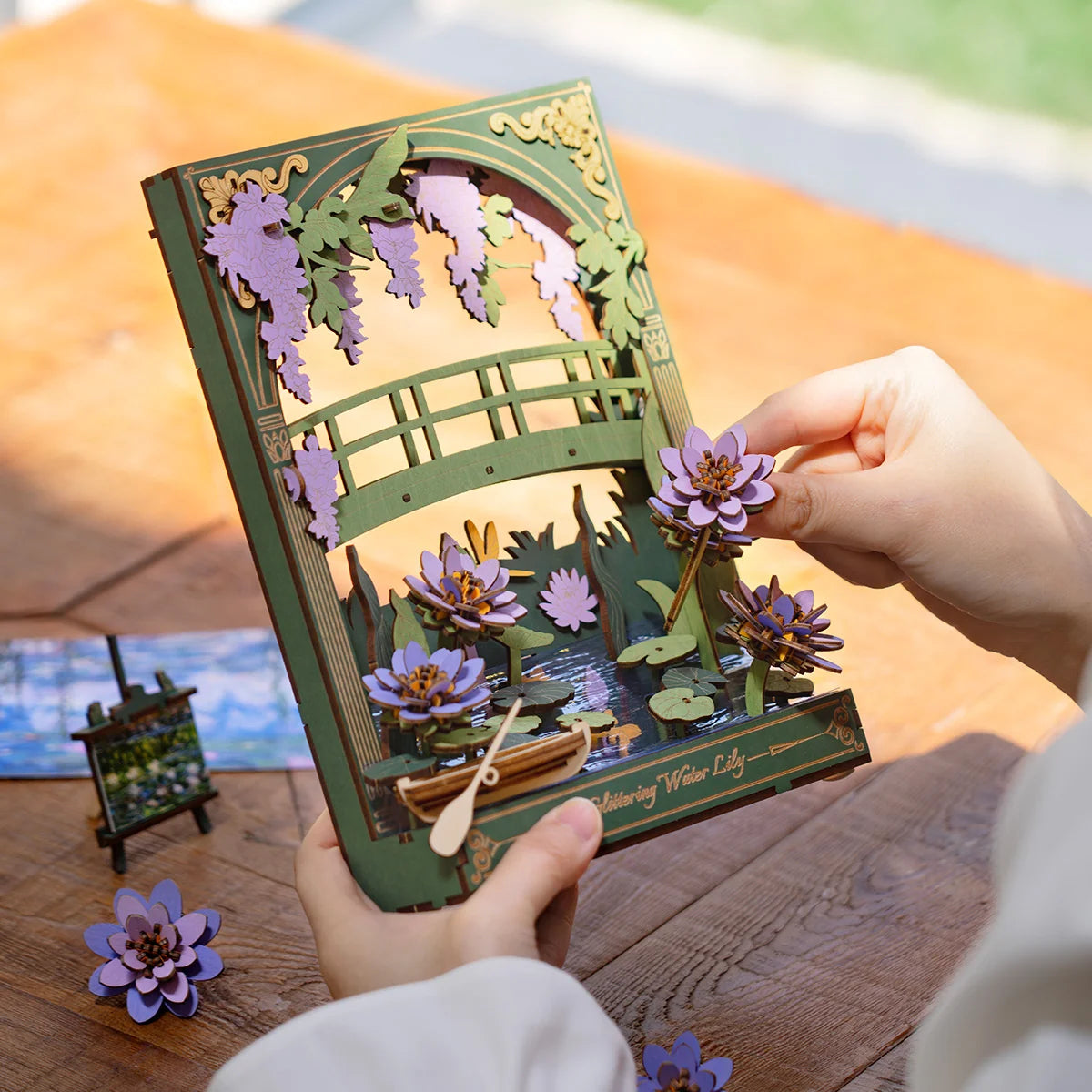 3D pop-up card with a bridge and flowers held by hands on a wooden surface.