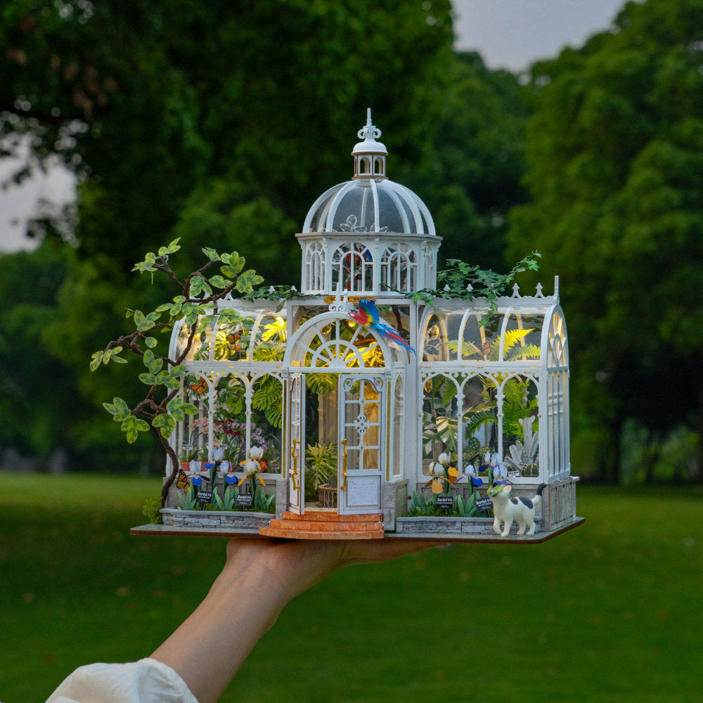 A miniature white glass conservatory model with warm yellow interior lights, small plants, birds, and a dog figurine, held up by a person's hand against a blurred green park background.