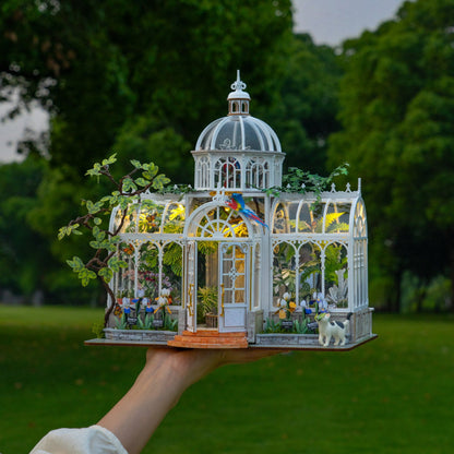 A miniature white glass conservatory model with warm yellow interior lights, small plants, birds, and a dog figurine, held up by a person's hand against a blurred green park background.