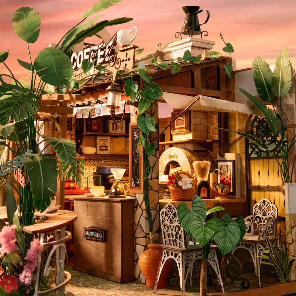 A highly detailed miniature diorama of a cozy, rustic Italian-style coffee shop and pizza oven, decorated with lush green plants, white metal chairs, and warm glowing lights under a pinkish sky.
