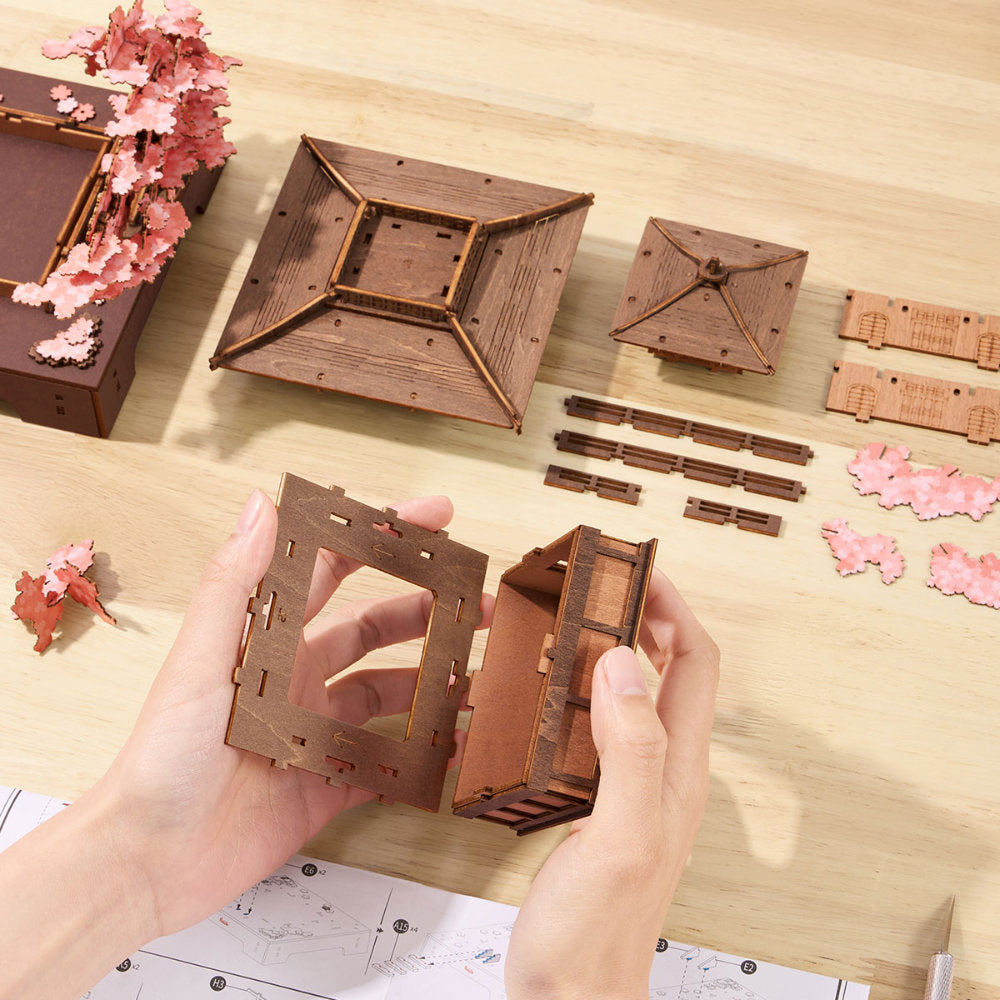 A pair of hands assembling a wooden 3D model of a traditional Japanese pagoda on a light wood tabletop, with other pre-cut wooden pieces and decorative pink cherry blossom ornaments arranged around it.